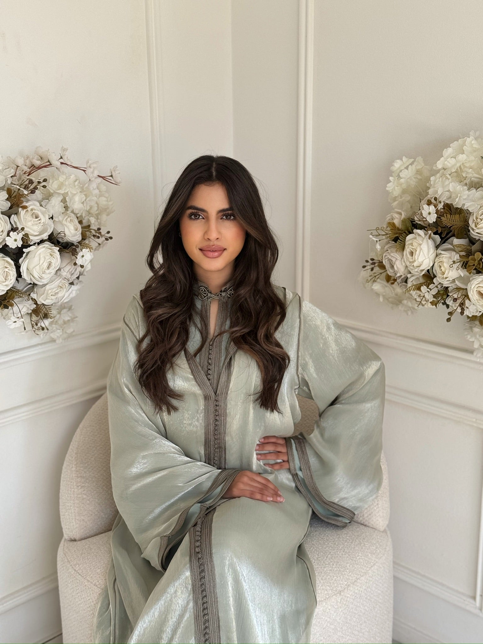Model wearing a silver-green Moroccan jellaba sitting with floral décor in a white interior