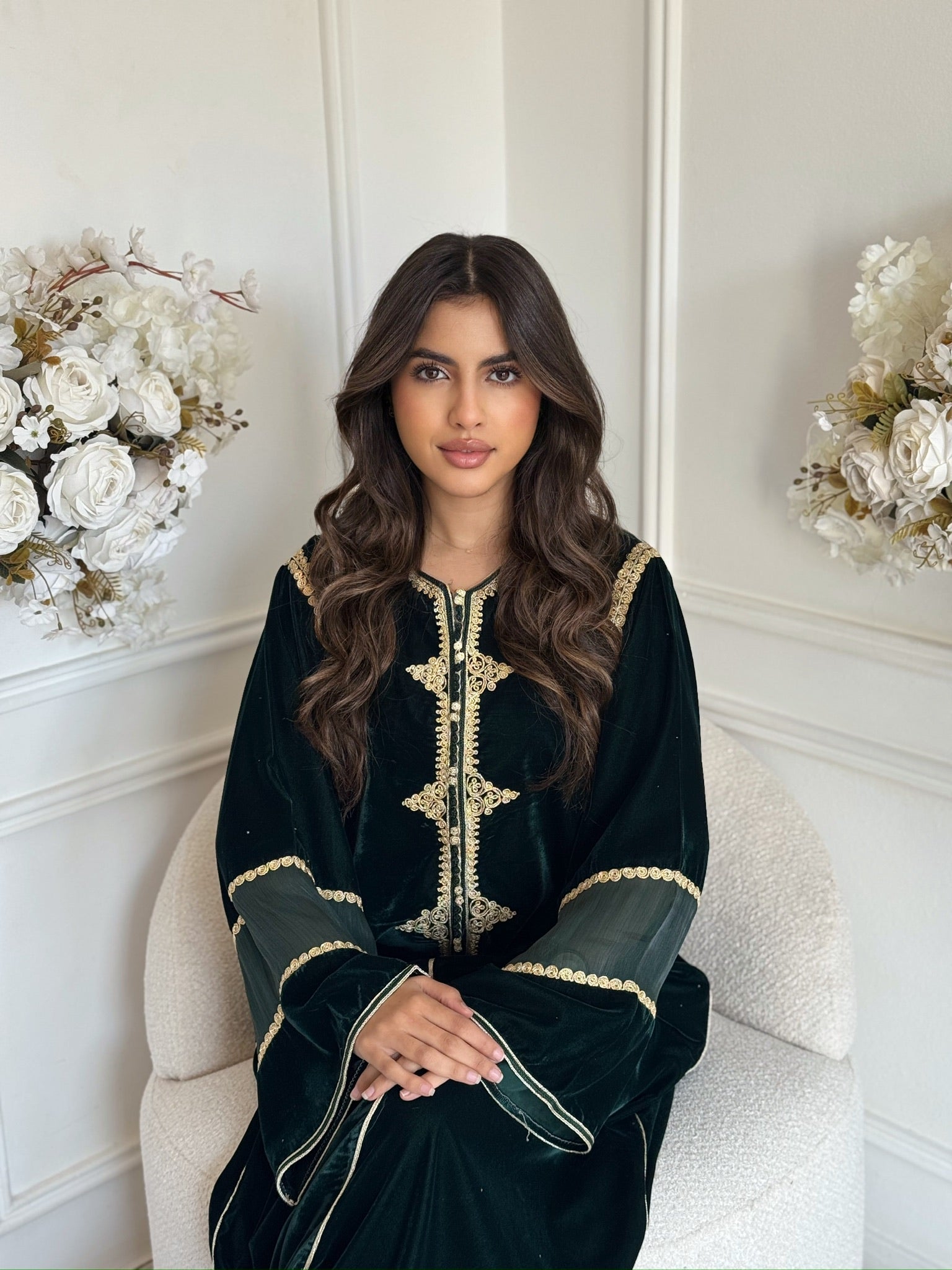 vModel wearing a black Moroccan gandoura with gold embroidery sitting indoors with white flowers
