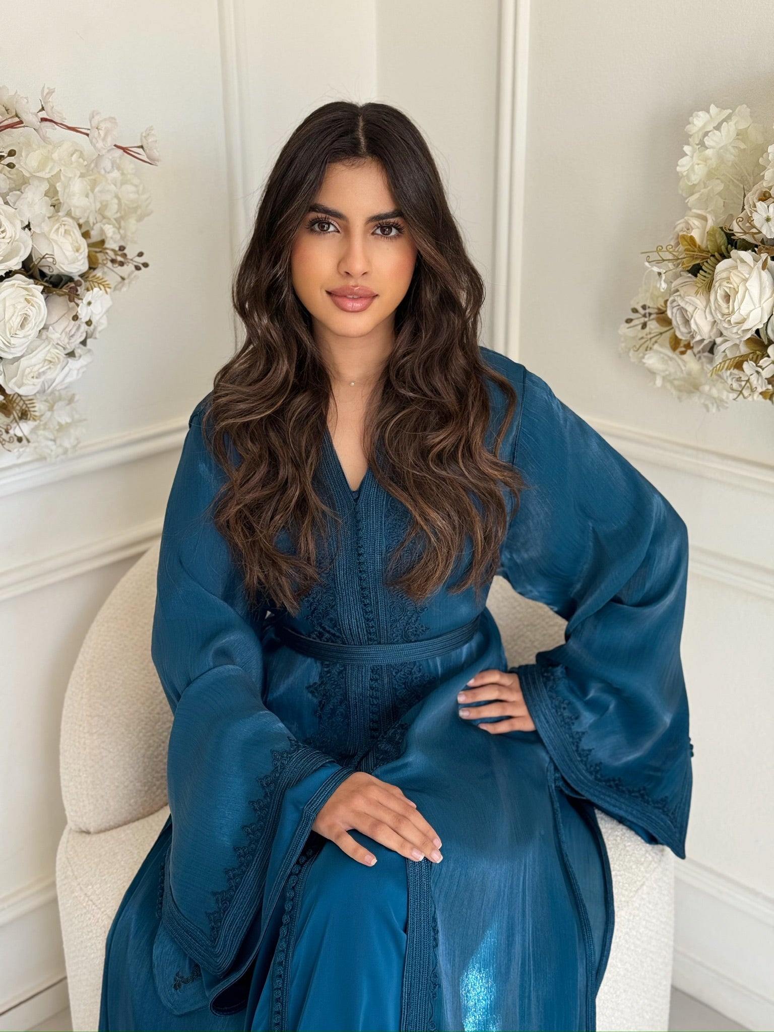 Model wearing a deep blue Moroccan takchita sitting with floral background in a white room