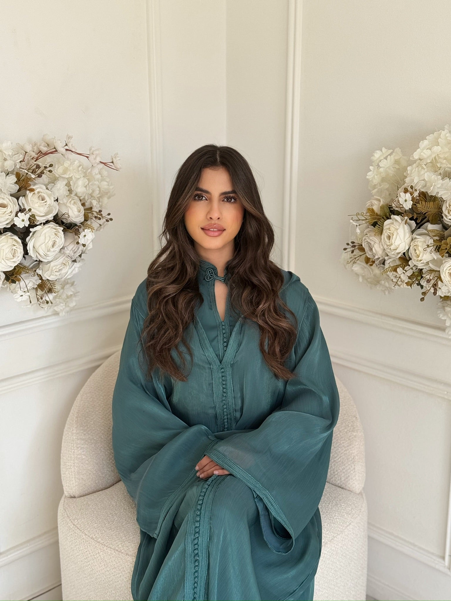 Model wearing a teal blue Moroccan jellaba sitting in a white room with floral décor